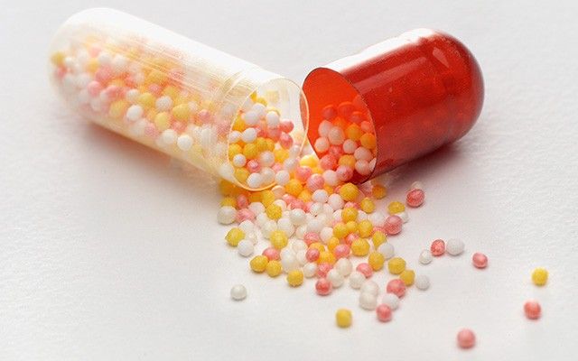 Eine geöffnete rot-transparente Kapsel liegt auf einer weißen Oberfläche, aus der viele kleine, bunte Kügelchen herausrollen.