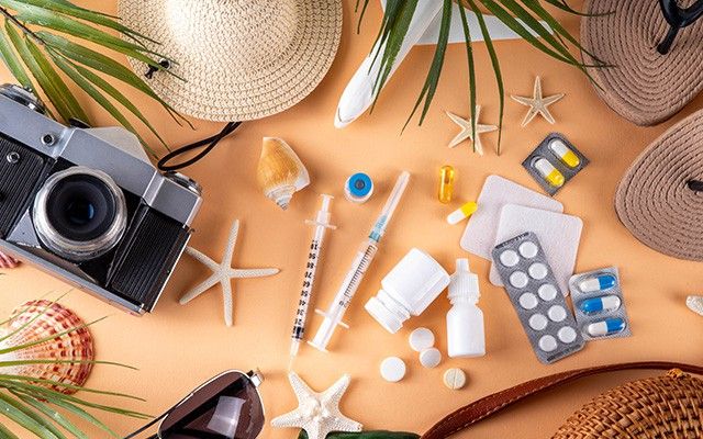 Verschiedene Medikamente wie Tablettenblister, eine Salbentube und Asthmasprays liegen dekorativ im Sand neben Muscheln, Seesternen und einer Strohtasche.