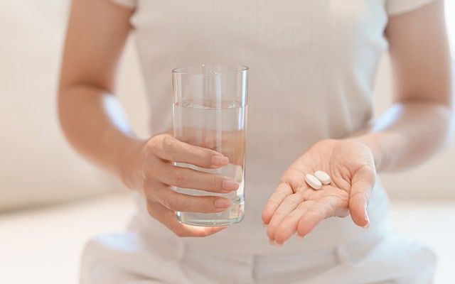 Eine Person hält in einer Hand ein Glas Wasser und zeigt auf der geöffneten Handfläche der anderen Hand zwei weiße Tabletten.
