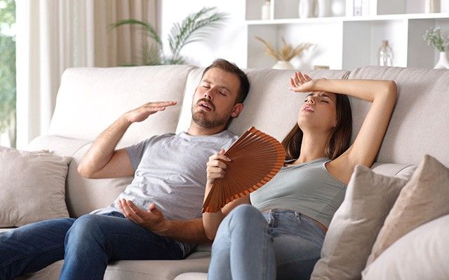 Ein Mann und eine Frau sitzen erschöpft auf einem Sofa im Wohnzimmer und fächeln sich bei Hitze Luft mit Zeitschriften zu.