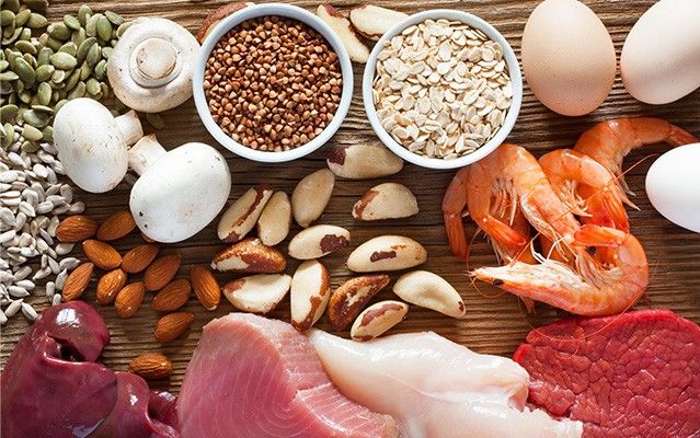 Eine Auswahl an selenreichen Lebensmitteln wie Nüsse, Pilze, Getreidekörner, Eier, rohes Fleisch und Garnelen auf einer dunklen Holzoberfläche.
