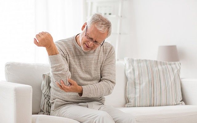 Ein älterer Mann sitzt auf einem hellen Sofa und reibt oder massiert sich mit besorgtem Gesichtsausdruck den linken Unterarm.