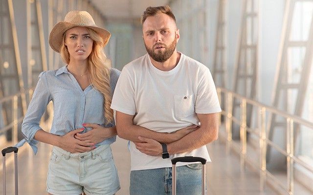 Ein Paar mit Koffern steht im Urlaub im Freien; beide blicken unwohl und halten sich mit schmerzverzerrtem Gesicht den Bauch.