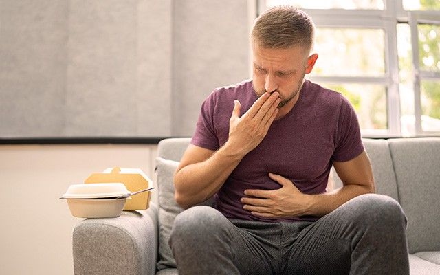 Ein Mann sitzt mit schmerzverzerrtem Gesicht auf einem Sofa und hält eine Hand an seinen Hals sowie die andere an den Magen.