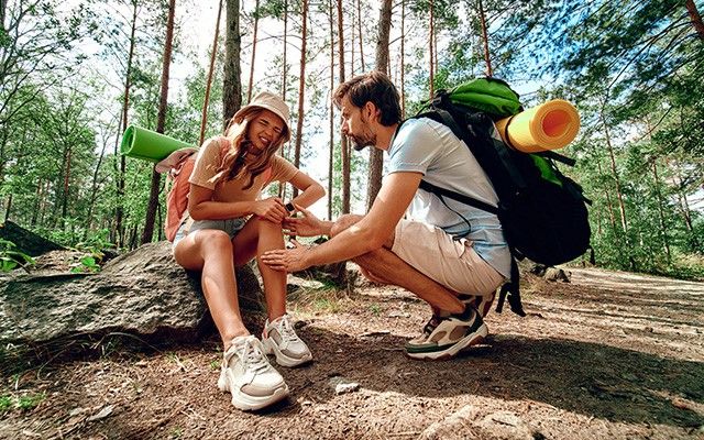 Zwei Wanderer mit Rucksäcken sitzen auf einem Felsen im Wald, während ein Mann einer Frau ein Pflaster auf das Knie klebt.
