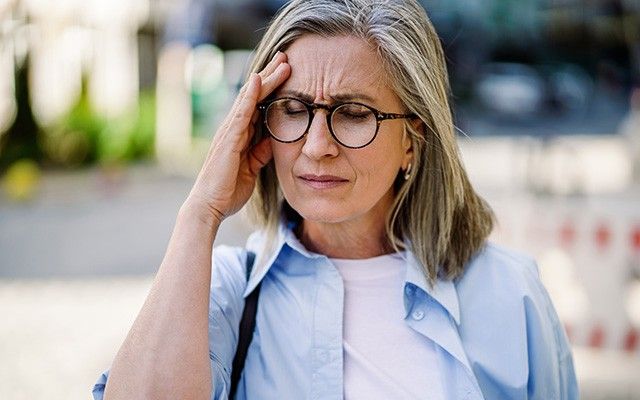 Eine ältere Frau mit Brille kneift im Freien schmerzerfüllt die Augen zusammen und hält sich eine Hand an die Schläfe.